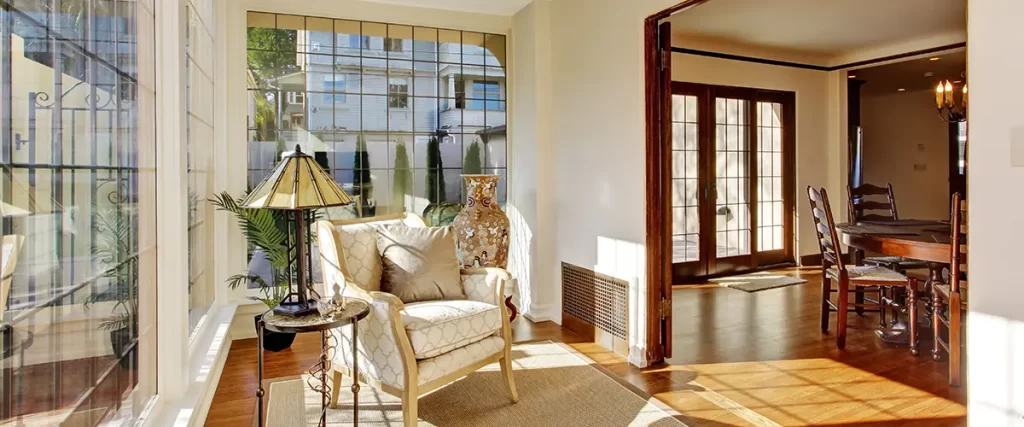 Bright sunroom with large windows, hardwood floors and comfortable seating beside a dining room.