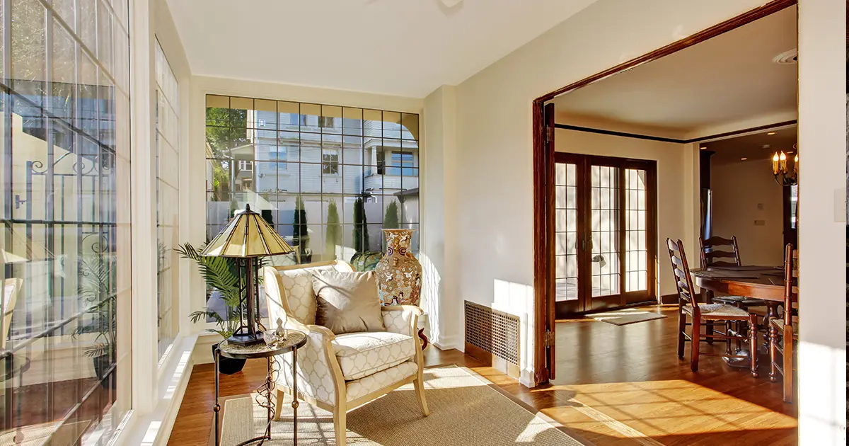 Bright sunroom with large windows, hardwood floors and comfortable seating beside a dining room.