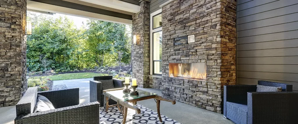 Covered outdoor living space with stone fireplace, wicker seating and modern home exterior.