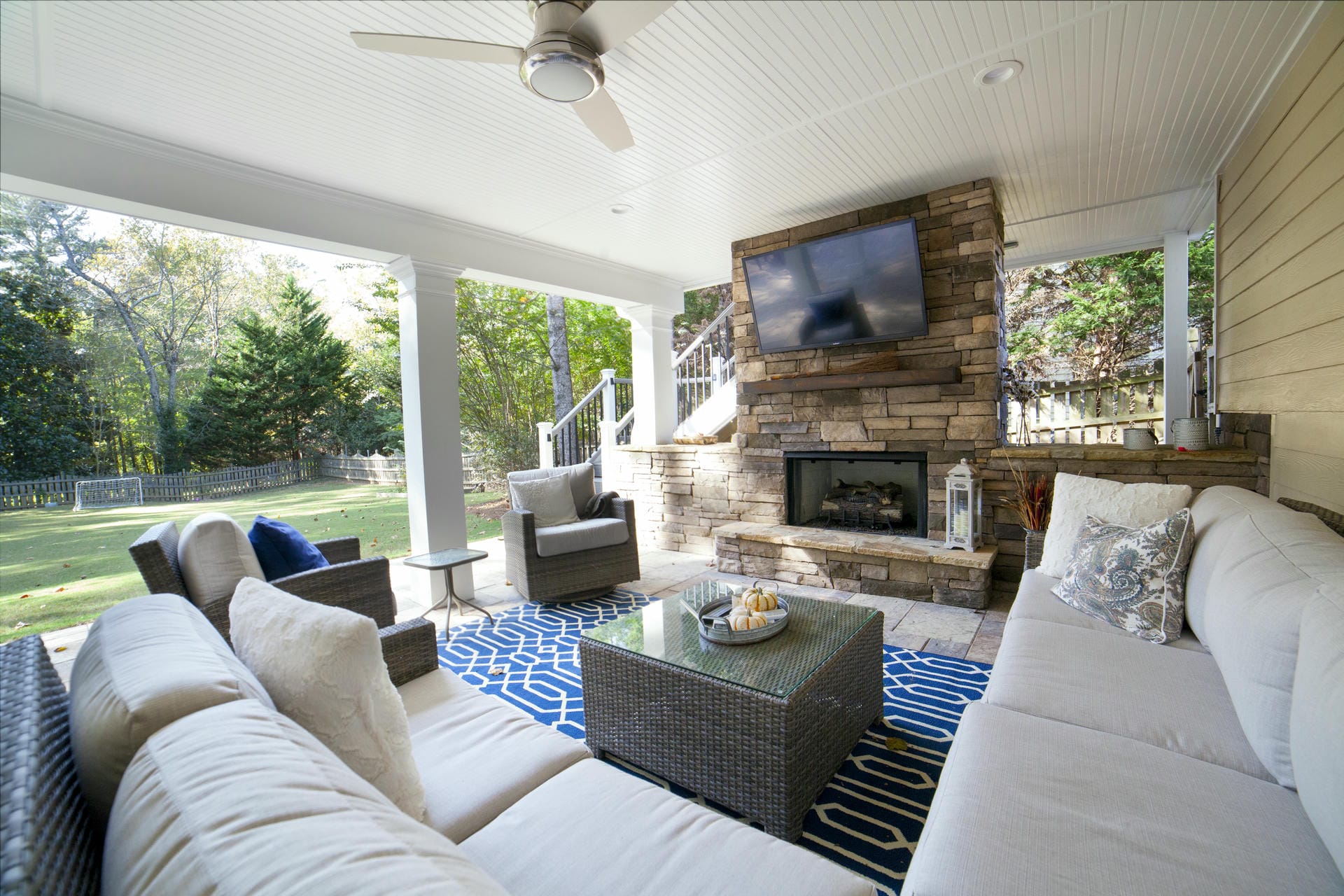 Covered outdoor living space with a stacked stone fireplace, mounted television, ceiling fan and wicker seating overlooking a fenced back garden