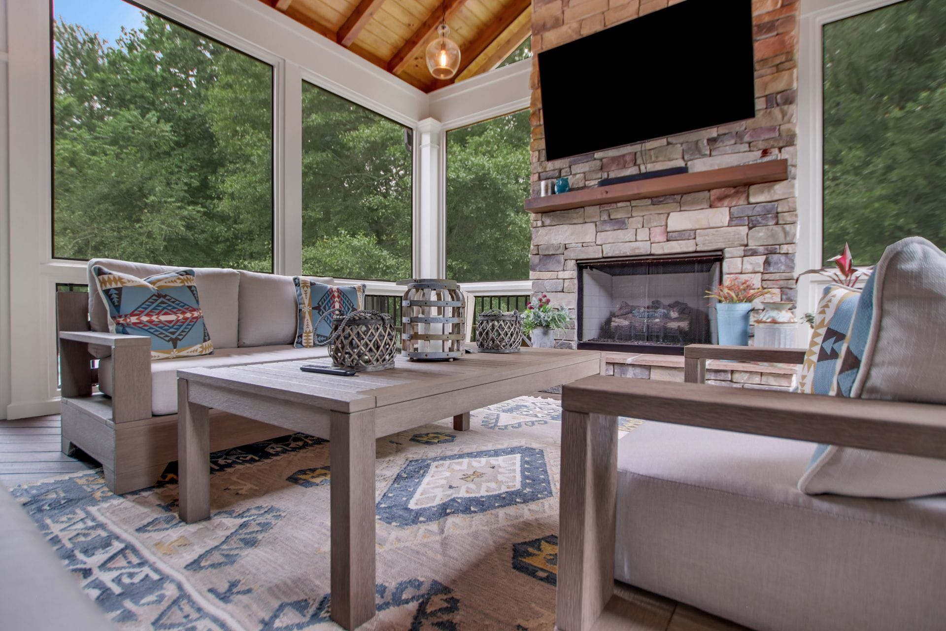 Screened porch furnished as a living room, with a stacked stone fireplace, a mounted television and a stained cedar rafter ceiling