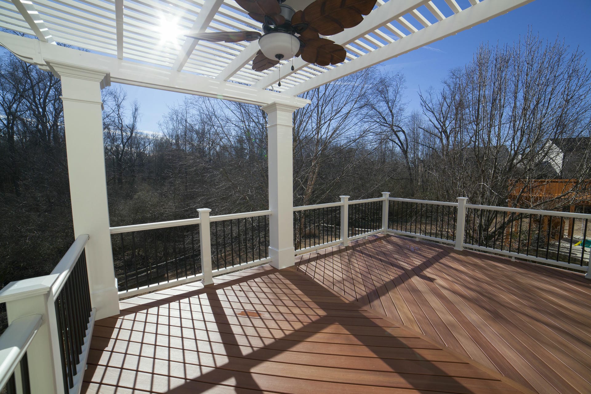 Composite deck beneath a white pergola with a ceiling fan and a wooded view
